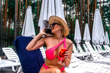 Güneş yatağında oturmuş, telefondan konuşurken gülümseyen bir kadın kokteyl ile güneşleniyor. Mutluluk duygusu, yaz tatili konsepti.