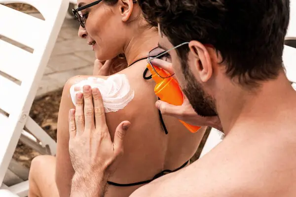 Man squeezing a sunscreen lotion out of the pump bottle on the back skin of his contented female companion sitting in front of him