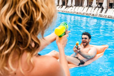 Sarışın kadın, güneş gözlüklü neşeli bir delikanlıyla su tabancası oynuyor. Açık havuzda şişme yatakta yatıyor.