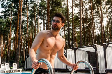 Güneşli bir günde havuzda poz veren kaslı, güneş gözlüğü takan mutlu adam. Yaz zamanı, tatiller, yaşam tarzı konsepti