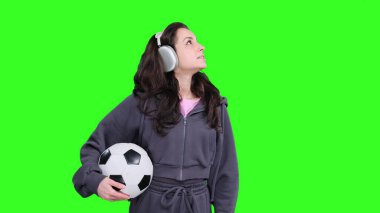 Mutlu kadın, kulaklık takıp yeşil alanda futbol topunu tutarken yukarı bakıyor.