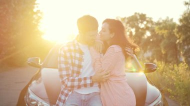 Romantik genç çift kucaklaşırken arabanın kaputuna yaslanıyor. Ulaşım, seyahat, ilişki, yaşam tarzı konsepti. Yavaş çekim