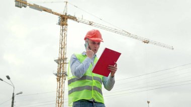 Yelekli ustabaşı, koruyucu miğfer yerinde duruyor, panoyu tutuyor ve telefonla konuşuyor. Yavaş çekim fotoğrafı