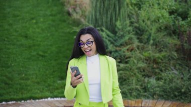 Yeşil ofis kıyafetleri içinde, elinde akıllı telefon olan ve kazanmaktan zevk alan, parkta yürüyen bir kadın. Yavaş çekim