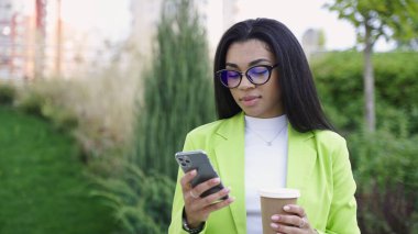 Yeşil ofis elbiseli bir kadın, elinde akıllı telefon ve sohbet, parkta durmuş, elinde kahve tutuyor. Yavaş çekim