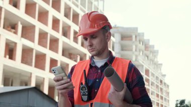 Koruyucu giysili ve miğferli bir adam binanın yanında duruyor, elinde planlar ve akıllı telefondan daktilo ediyor. Yavaş çekim