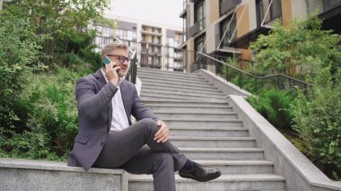 Elinde cep telefonu olan bir iş adamı dışarıda merdivenlerin yanında otururken konuşuyor. İş, iletişim, teknoloji konsepti. Yavaş çekim