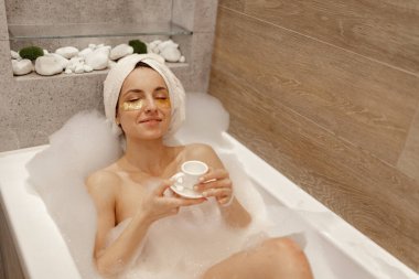 Mutlu, çıplak, güzel bir kadın. Gözleri kapalı, kafasında havluyla küvette uzanmış. Elinde kahve fincanıyla dinleniyor. Hijyen ve vücut yıkama konsepti