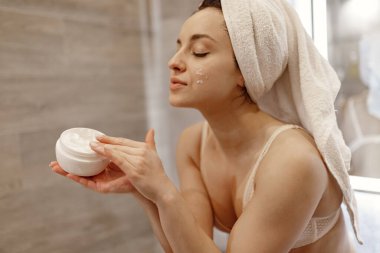 İç çamaşırlı güzel bir kadın banyoda elinde vücut losyonu tutarken beyaz havluyla poz veriyor. Cilt tedavisi kavramı