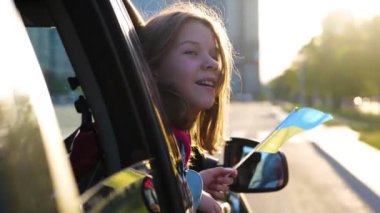 Little Caucasian girl in car window with Ukrainian flag in hand. Child smiles at wind in vehicle window. Pretty small female kid holding flag of Ukraine looks out of automobile. Family trip. Close up