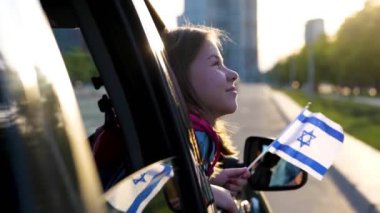 İsrail bayrağını elinde tutan neşeli küçük kız araba camından dışarı bakıyor. İsrail 'de sevimli şirin çocuk seyahatleri. Vatansever ruh hali. Aile gezisi. Seyahat konsepti. Gerçek zamanlı video