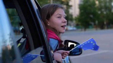 Close up of Caucasian pretty kid looking out car window with flag of European Union in hand. Small girl holds EU flag sitting in vehicle on family trip. Travel in automobile in Europe, journey concept