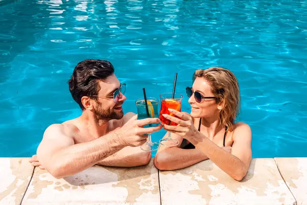 stock image Smiling young woman and a cute dark-haired guy clinking glasses of cocktails in an outdoor swimming pool. Leisure and summer vacation concept