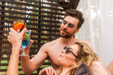 Blonde young woman and her boyfriend clinking glasses with cocktails while resting in a gazebo. Romance and summer vacation concept clipart