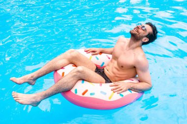 Mutlu genç adam yüzme havuzunda dinlenirken gözlerini kapatıyor, şişme halkanın üzerinde oturuyor. Yaz zamanı, tatiller, yaşam tarzı konsepti