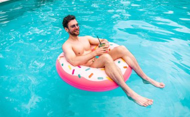 Güneş gözlüklü mutlu atletik adam yüzme havuzunda eğleniyor, elinde kokteyl var. Yaz zamanı, tatiller, yaşam tarzı konsepti