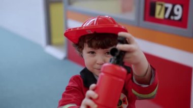 Gülümseyen itfaiyeci çocuk elinde oyuncak söndürücü, üniforma giyiyor, oyun merkezinde oynuyor. Ağır çekim. Çocukluk, eğlence, meslek kavramı