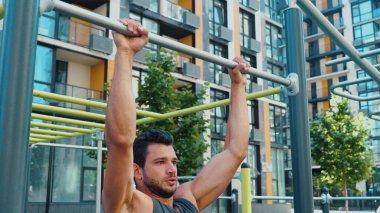 Genç adam dışarıda egzersiz yapıyor. Kamera güçlü bir sporcunun barfiks çekmesinden sonra hareket ediyor. Modern kentsel binalarda spor faaliyetleri ve spor faaliyetleri