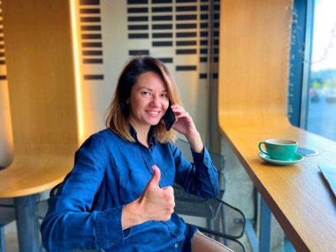 Kafkasyalı neşeli pozitif kadın cep telefonuyla konuşuyor kafeteryada oturmuş kameraya bakıyor ve gülümsüyor. Akıllı telefon iletişimi. Portre fotoğrafı. Kadın cep telefonu kullanıyor.