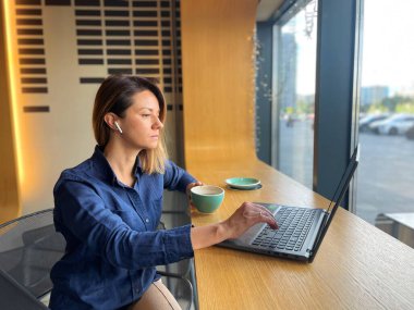 Kablosuz kulaklıklı meşgul beyaz iş kadını portresi bir fincan kahveyle kafede oturuyor ve internette dizüstü bilgisayar sörfü yapıyor. Kadın bilgisayarda daktilo ediyor ve kahve içiyor.