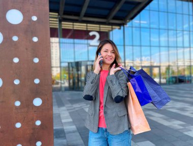 Alış veriş yapan beyaz, mutlu, güzel bir kadın. Açık havada dururken akıllı telefondan konuşuyor. Neşeli, pozitif, yetişkin bir kadın elinde bir sürü kese kâğıdıyla cep telefonundan arıyor. Konsept satın alınıyor