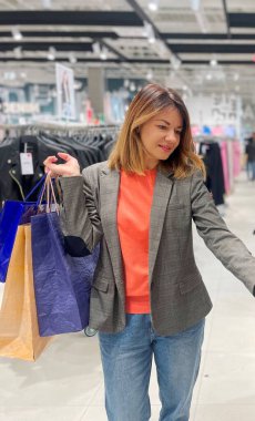 Alışveriş sırasında mağazada duran elbiselere bakan mutlu beyaz kadın. Neşeli, olgun bir bayan alıcı alışveriş merkezinden bir şeyler alıyor. Kıyafet mağazası. Dikey çekim. Sezon satışı konsepti