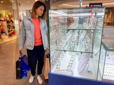 Kot ceketli güzel beyaz bir kadın alışveriş merkezinde elinde bir sürü paketle lüks mücevherlerle vitrinlere bakıyor. Mağazada mücevher seçen bir kadın. Satışlar