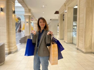 Alışveriş merkezinin içinde elinde bir sürü kese kağıdıyla tek başına duran günlük kıyafetlerle çekici bir yetişkin kadın. Mutlu kadın alışveriş torbaları dolusu parayı kameraya bakarak harcadı. Alışveriş çılgınlığı.