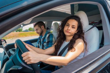 Erkek eğitmen sınav raporunda notlar alırken, sevimli bir bayan araba kullanmak için ders çalışıyor. Ehliyet sınavı, ehliyet kursu, sınav konsepti.