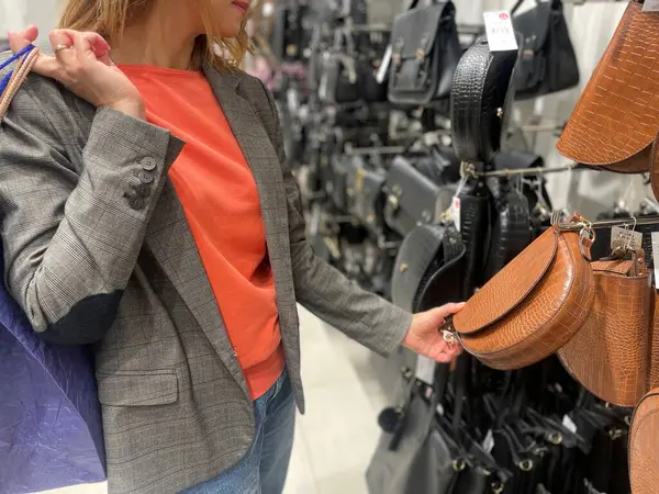 Mağazada durup kahverengi deri çantaya bakan şık bir kadın. Alışveriş aktivitesi. Genç beyaz kadın alışveriş merkezinden çanta aksesuarları alıyor. Moda mağazası, sezon satış konsepti.