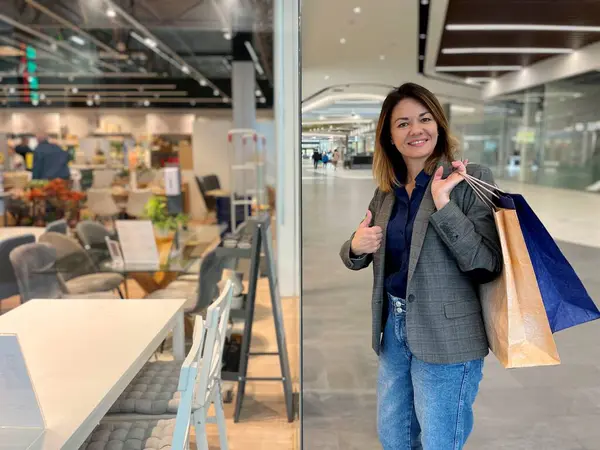 Alışveriş torbaları tutan ve kameraya bakıp gülümseyen güzel bir kadın alışveriş merkezinin yanında elinde mobilyalarla başparmağıyla dikiliyor. Yemek odası mobilyası, alıcı konsepti.