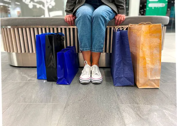 Şehir merkezindeki kapalı bir bankta oturan tanınmayan bir kadın yerde bir sürü alışveriş torbası var. Alışveriş merkezindeki kağıt torbaların yanındaki kadın bacaklarını kapat. Satışlar, satın alma kavramı