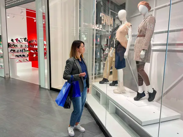 Beyaz tenli güzel bir kadın alışveriş merkezinde elinde çantalarla durmuş mankenlerle vitrinlere bakıyor. Moda kıyafetleriyle mağazanın yakınında alışveriş yapan güzel bir kadın. Alıcı konsepti