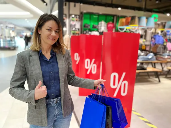 Alışveriş merkezinde elinde çantalarla durup kameraya bakan ve baş parmağını gösteren mutlu beyaz bir kadın. Satış mağazasının yanında güzel bir kadın alışverişi sever. Alıcı konsepti