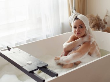 Küvette köpükle oturmuş vücudunu yıkayan ve keyifle kameraya bakan çekici genç bayan. Sabah hijyen rutini. Kafkasyalı mutlu, güzel bir kadın, kafasında havluyla banyo yapıyor.