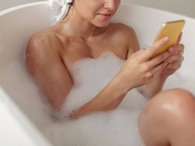 Kapatın. Beyaz, çekici, çıplak bir kadın küvette uzanıyor ve akıllı telefondan yazı yazıyor. Ev hanımı evde dinleniyor, banyo yapıyor ve internette cep telefonuna bakıyor. Banyo yaparken alet kullanan bir kadın.