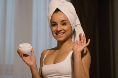 Close up of happy smiling beautiful young woman after shower with towel on head using face cream and looking at camera in positive mood. Beauty and wellness, female using skincare product at home