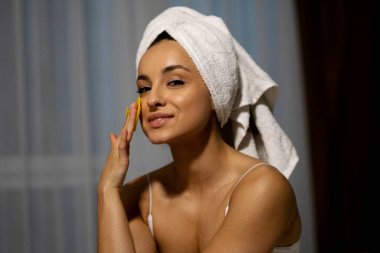 Close up of happy pretty young smiling woman with towel on head using facial toning device at home looking at camera. Beauty procedure. Beautiful lady using skin face massage item. Beauty device