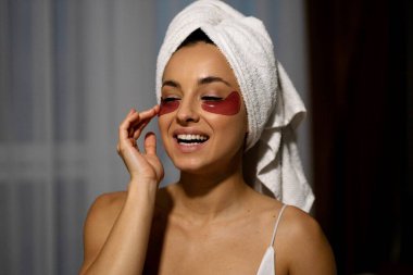 Caucasian joyful happy woman using facial cosmetic products at home and smiling. Close up of beautiful young female applying eye patches on face skin. Beauty treatment. Skincare. Skin care routine.