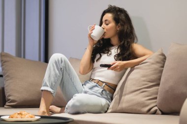 Beautiful Caucasian young woman sitting on sofa alone watching TV and drinking coffee. Attractive happy brunette female resting at home holding remote control in hand watching movie. Indoor concept
