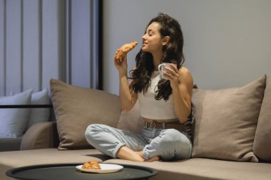 Beautiful Caucasian lady sitting on couch at home in good mood smelling fresh croissant and holding cup of coffee in hand. Pretty joyful female with dark hair enjoying morning coffee with croissant