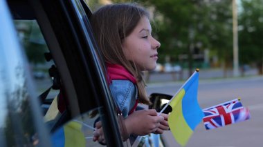 Ön koltukta oturan beyaz çocuk elinde İngiliz ve Ukrayna bayraklarıyla arabanın camından dışarı bakıyor. Ukrayna ve İngiltere 'nin bayrağı küçük kızda. Mülteciler savaştan kaçıyor. Konsepti kapat, otomatik seyahat et