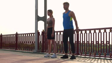 Camera approaching young couple of man and woman in sportswear working out outside on the bridge. Caucasian sporty male and female doing exercise outdoor. Athletes doing squats. Sport activity.
