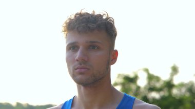 Close up of handsome Caucasian athletic male in sportswear standing outside and looking away. Young serious man stands outdoors in city on sky background. Sporty lifestyle. Sportsman concept
