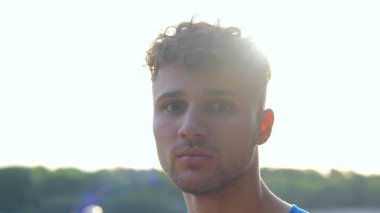 Close up of joyful handsome Caucasian male athlete in good mood standing outside and looking at camera. Young happy man stands outdoors in city smiling to camera. Sporty lifestyle. Sportsman concept