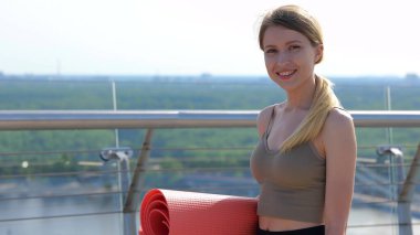 Close up portrait of beautiful young fit woman looking at camera and smiling in good mood before training outdoors. Pretty female with gym mat outside showing thumb up. Physical activity. Slow motion