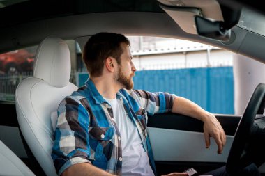 Genç sakallı adam arabada oturup cep telefonu tutarken pencereden dışarı bakmaktan zevk alıyor. Yan görüş. Yaşam tarzı, ulaşım kavramı