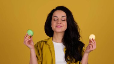 Gülümseyen bayan ısırırken makarondan zevk alıyor. Sarı arka planda duran kadın. Tatlı yemek konsepti. Gerçek zamanlı.