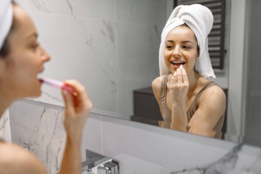 Gülümseyen güzel bayan banyoda aynanın önünde dikilirken dudaklarına çıplak ruj sürüyor. Makyaj, sabah ve yaşam tarzı rutini