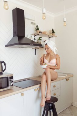 İç çamaşırlı, mutfak tezgahına oturmuş, elinde salata tabağıyla genç, beyaz bir kadın resmi. Evde kahvaltıda salata yiyen bir kadın. Sağlıklı gıda konsepti